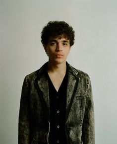 a young man standing in front of a white wall wearing a black shirt and jacket