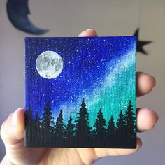 a hand holding up a small painting with trees and the moon in the background