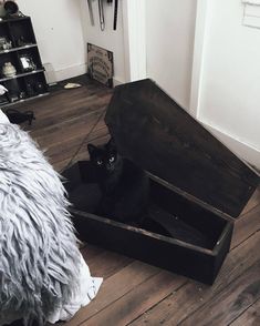 a black cat sitting in an open wooden box on the floor next to a bed