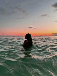 a woman sitting in the ocean at sunset with her back to the camera and looking out into the distance