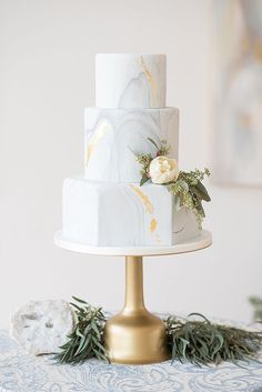 a white and gold wedding cake sitting on top of a metal stand with greenery