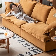 a woman laying on top of a brown couch reading a book in a living room