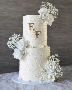 a three tiered wedding cake with white flowers and pearls on the top, decorated with monograms