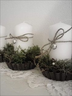 three white candles with moss in them on a lace tablecloth