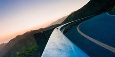 a curved road on the side of a mountain with mountains in the background at sunset