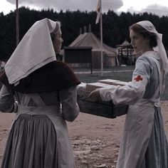 two women dressed in historical clothing carrying a wooden box with something on it's back