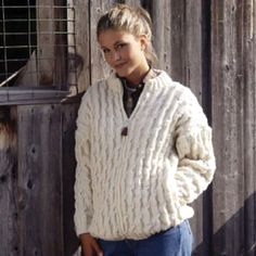 a woman standing in front of a wooden fence wearing a white sweater and blue jeans