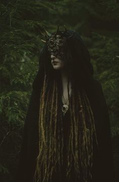 a woman with dreadlocks wearing a horned mask and black dress in the woods