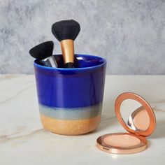 a blue cup with makeup brushes in it next to a compact mirror on a marble surface