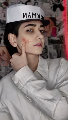 a woman with makeup on her face wearing a baseball cap and white shirt, standing in front of a wall