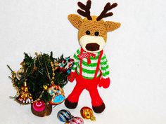 a knitted reindeer standing next to a small christmas tree with ornaments around it on a white surface