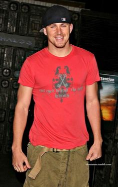 a man in a red shirt and hat smiles at the camera while standing next to a wall