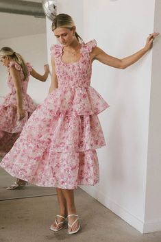 The Absedee Floral Midi Dress is crafted with a smocked bodice that complements its scoop neckline. It boasts charming ruffled cap sleeves and a gracefully flowing three-tiered skirt. The dress is designed with a printed tulle shell that adds a delicate touch, and it is fully lined to ensure both comfort and coverage. Knee-length Smocked Dress With Ruffles For Garden Party, Fitted Smock Tiered Midi Dress, Fitted Tiered Smock Midi Dress, Fitted Tiered Midi Dress With Smock Detail, Pink Fitted Tiered Smocked Dress, Smocked Tiered Skirt Dresses For Garden Party, Fitted Pink Tiered Smocked Dress, Smocked Tiered Skirt Dress For Daywear, Fitted Smock Dress With Tiered Skirt