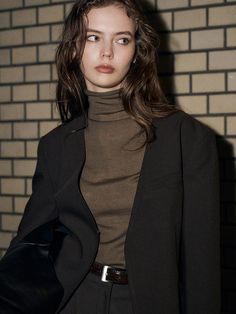 a woman in a brown turtle neck sweater and black pants standing next to a brick wall