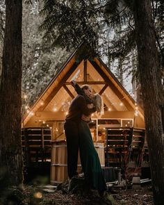 a man and woman standing in front of a wooden cabin surrounded by trees with lights