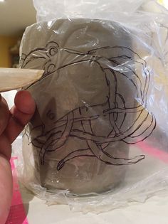 a person holding a spoon in front of a cup with drawings on it and plastic wrap