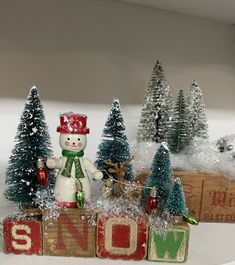 a small snowman sitting on top of a wooden block in front of some christmas trees