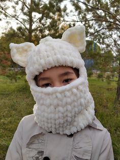 a young boy wearing a knitted animal mask and jacket in the grass with trees behind him