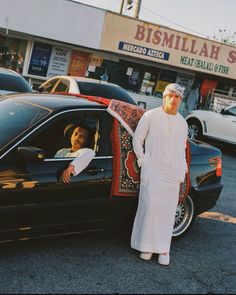 a man standing next to a black car in front of a building with a woman sitting in it