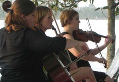 three women are playing violin in front of a lake with trees and water behind them