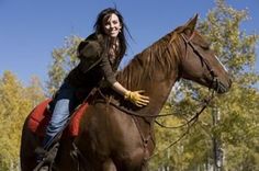 a woman riding on the back of a brown horse next to trees and blue sky