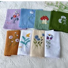 six embroidered napkins with flowers on them sitting on a white tablecloth next to a bouquet of wildflowers