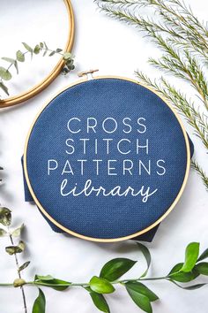 the cross stitch pattern is shown on a blue hoop with greenery next to it