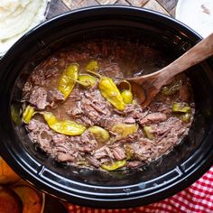 a crock pot filled with beef and peppers next to potatoes on a wooden table