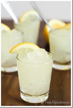 three glasses filled with lemonade and ice on top of a wooden table next to spoons