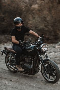 a man riding on the back of a black motorcycle down a dirt road next to a forest