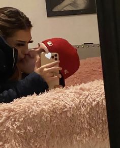 a woman laying on top of a bed holding a cell phone to her face while wearing a red hat