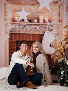 a man and woman sitting next to each other in front of a fireplace with christmas decorations