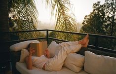 a person laying on top of a white couch next to a palm tree and reading a book