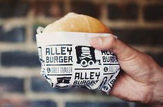 a person holding up a sandwich in front of a brick wall with the words alley burger written on it