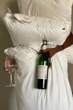 a person holding a bottle of wine on top of a bed with white sheets and pillows