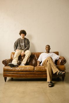 two men sitting on top of a brown leather couch next to each other in front of a white wall