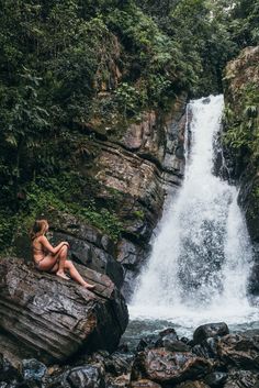 a woman sitting on top of a large rock next to a waterfall in the forest