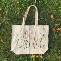 a white tote bag sitting on top of a green grass covered field with flowers