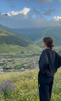 a woman standing on top of a lush green hillside next to a valley filled with flowers