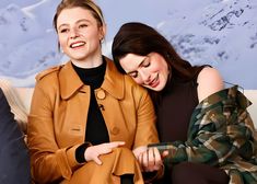 two women sitting on a couch with one pointing at her phone and the other smiling