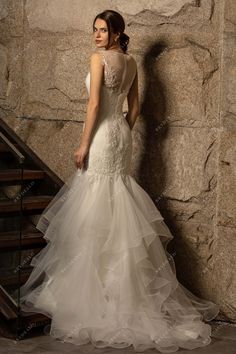 a woman standing in front of a stone wall wearing a wedding dress with ruffled layers