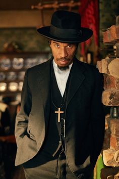 a man wearing a black suit and hat with a cross on the chest standing next to a brick wall