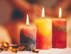 three different colored candles sitting next to each other on a white tablecloth with someone's hand in the background