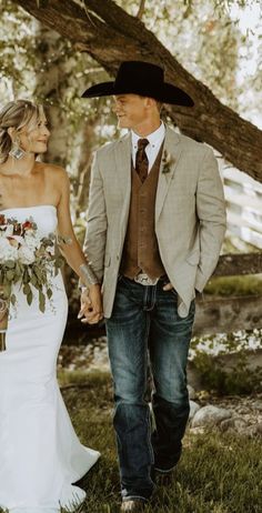 a bride and groom holding hands walking through the grass under a tree with their cowboy hats on