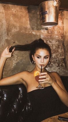 a woman sitting in a chair holding a drink and looking at the camera with one hand on her head