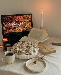 an open book on a table next to a cake and coffee cup with a laptop in the background