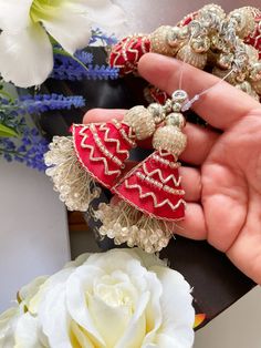 a hand holding two red and gold earrings in front of some white flowers on a table