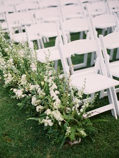 rows of white folding chairs with flowers on the grass in front of them at an outdoor wedding ceremony