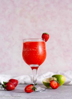 a strawberry margarita in a wine glass with limes and strawberries around it