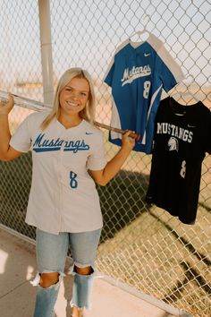 a woman holding a baseball bat in front of a fence with two shirts hanging on it